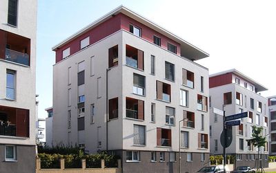 Ein puristischer, von Immobilienbewertern sachkundig überprüfter, Neubau eines Wohnhausblocks ist architektonisch einem Karton mit angehobenem Deckel nachempfunden. Die kubische Architektur wird durch asymmetrische Anordnung von Fenstern aufgelockert.
